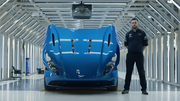 Todd Downs: male manufacturing engineering apprentice in dark coloured uniform standing to the side of a blue Aston Martin with its bonnet open