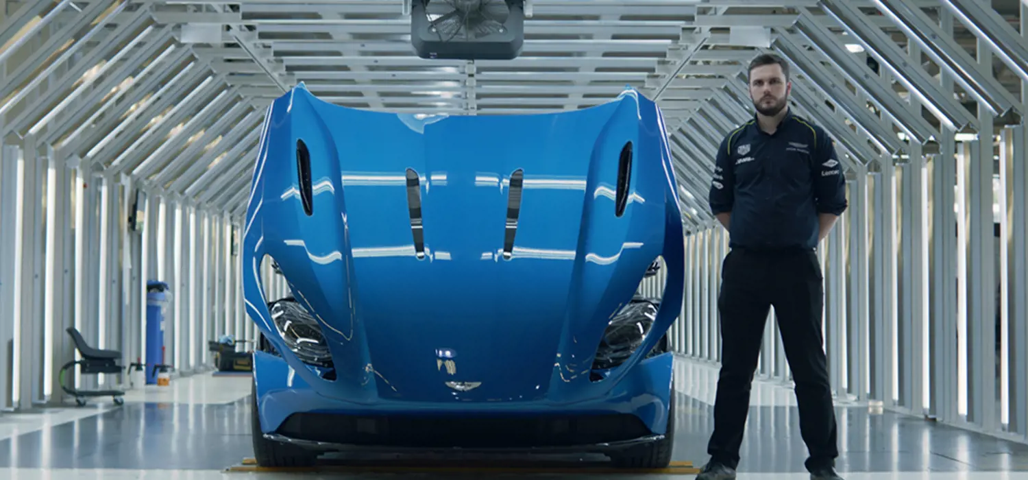 Todd Downs: male manufacturing engineering apprentice in dark coloured uniform standing to the side of a blue Aston Martin with its bonnet open