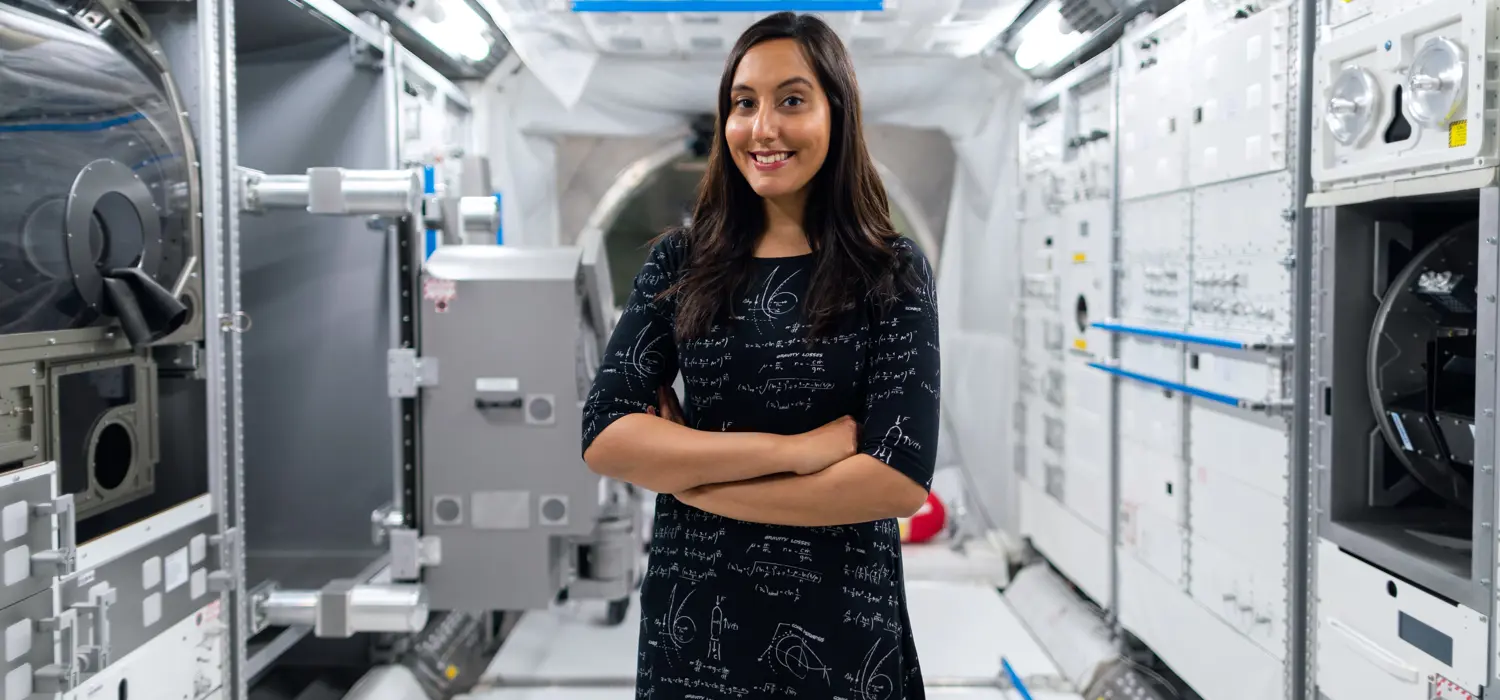 Vinita Marwaha Madill standing and smiling with her arms crossed with a background of space technology equipment.