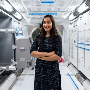Vinita Marwaha Madill standing and smiling with her arms crossed with a background of space technology equipment.